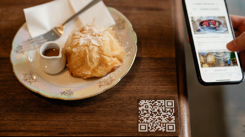 QR kód na stole vedľa dezertu na tanieri s karamelovou omáčkou a vidličkou, zobrazený vedľa mobilného telefónu, ktorý zobrazuje digitálne menu.